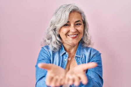 Foto de Mujer de mediana edad con el pelo gris de pie sobre fondo rosa sonriendo con las palmas de las manos juntas recibiendo o dando gesto. retención y protección - Imagen libre de derechos
