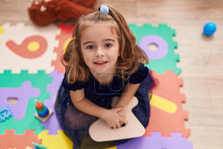 Foto de Adorable chica hispana sonriendo confiada sentada en el suelo en el jardín de infantes - Imagen libre de derechos