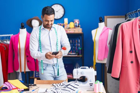 Photo for Young hispanic man tailor smiling confident using smartphone at sewing studio - Royalty Free Image