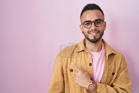 Téléchargez les photos : Jeune homme hispanique debout sur fond rose gai avec un sourire du visage pointant avec la main et le doigt sur le côté avec une expression heureuse et naturelle sur le visage - en image libre de droit