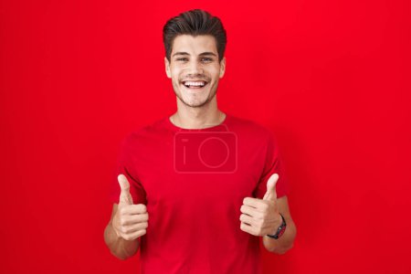 Téléchargez les photos : Jeune homme hispanique debout sur fond rouge signe de succès faisant geste positif avec la main, pouces levés souriant et heureux. expression joyeuse et geste gagnant. - en image libre de droit