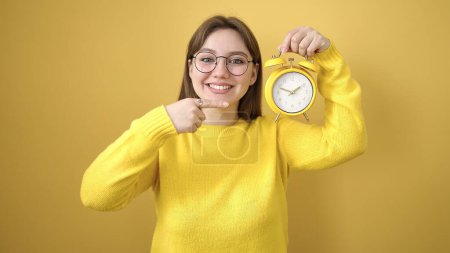 Photo for Young blonde woman smiling confident pointing with finger to alarm clock over isolated yellow background - Royalty Free Image