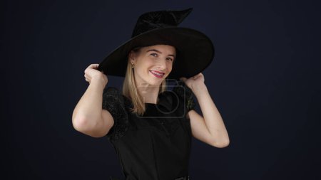 Foto de Mujer rubia joven con traje de bruja sosteniendo sombrero sobre fondo negro aislado - Imagen libre de derechos