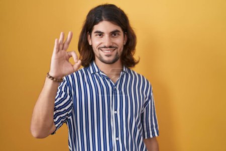 Photo for Hispanic man with long hair standing over yellow background smiling positive doing ok sign with hand and fingers. successful expression. - Royalty Free Image