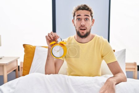Photo for Hispanic man with beard holding alarm clock in the bed scared and amazed with open mouth for surprise, disbelief face - Royalty Free Image