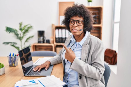Foto de Mujer negra con el pelo rizado usando auriculares de agente de call center en la oficina señalando a un lado preocupado y nervioso con el dedo índice, preocupado y expresión sorprendida - Imagen libre de derechos