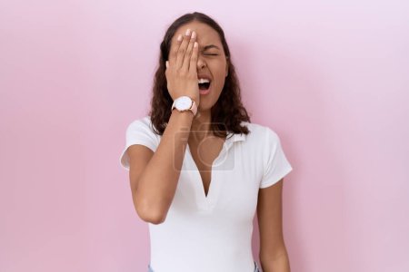 Téléchargez les photos : Jeune femme hispanique portant un t-shirt blanc décontracté bâillant fatigué couvrant la moitié du visage, des yeux et de la bouche avec la main. mal au visage dans la douleur. - en image libre de droit