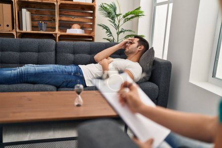Foto de Hombre hispano joven estresado teniendo terapia mental tumbado en sofá en el centro de psicología - Imagen libre de derechos