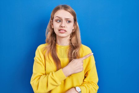 Téléchargez les photos : Jeune femme caucasienne debout sur fond bleu pointant de côté inquiet et nerveux avec index, expression inquiète et surprise - en image libre de droit