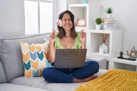 Foto de Hispanic young woman using laptop at home gesturing finger crossed smiling with hope and eyes closed. luck and superstitious concept. - Imagen libre de derechos