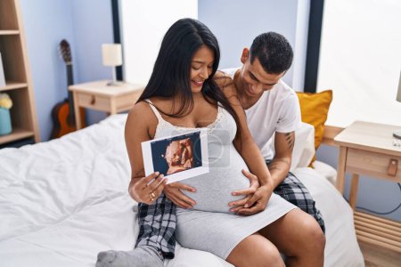 Photo for Young latin couple expecting baby hugging each other holding ecography at bedroom - Royalty Free Image