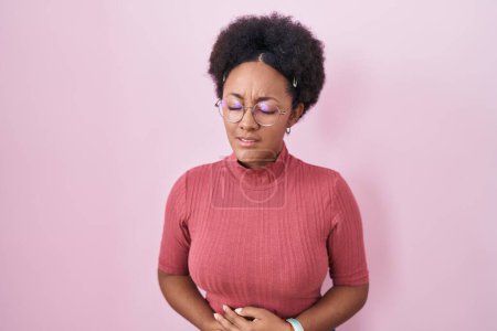 Foto de Beautiful african woman with curly hair standing over pink background with hand on stomach because indigestion, painful illness feeling unwell. ache concept. - Imagen libre de derechos