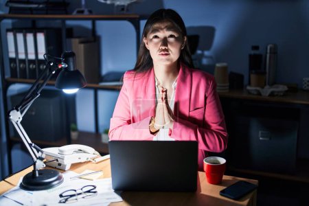 Téléchargez les photos : Jeune femme chinoise travaillant au bureau la nuit suppliant et priant avec les mains ainsi que l'expression de l'espoir sur le visage très émotionnel et inquiet. mendier. - en image libre de droit