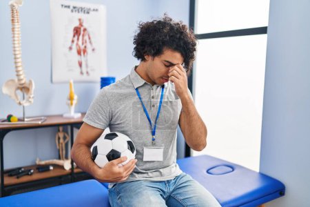 Foto de Hispanic man with curly hair working as football physiotherapist tired rubbing nose and eyes feeling fatigue and headache. stress and frustration concept. - Imagen libre de derechos