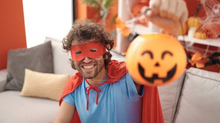 Téléchargez les photos : Young hispanic man wearing superhero costume holding halloween pumpkin basket at home - en image libre de droit