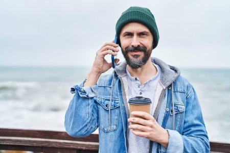 Foto de Joven hombre calvo hablando en el teléfono inteligente beber café en la playa - Imagen libre de derechos