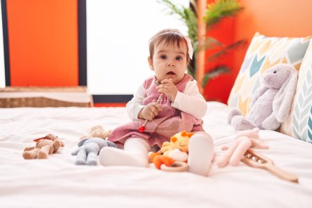 Foto de Adorable bebé hispano jugando maraca sentado en la cama en el dormitorio - Imagen libre de derechos