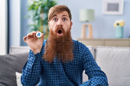 Foto de Caucasian man with long beard holding virtual currency bitcoin scared and amazed with open mouth for surprise, disbelief face - Imagen libre de derechos