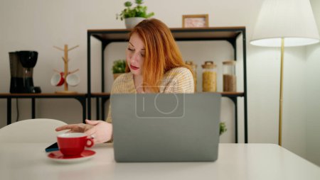 Foto de Joven pelirroja usando laptop bebiendo café en casa - Imagen libre de derechos