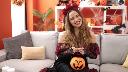 Foto de Joven hermosa mujer hispana con traje de katrina sosteniendo cesta de calabaza de Halloween en casa - Imagen libre de derechos