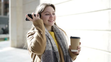Téléchargez les photos : Jeune femme blonde écoutant un message audio par le smartphone boire du café dans la rue - en image libre de droit