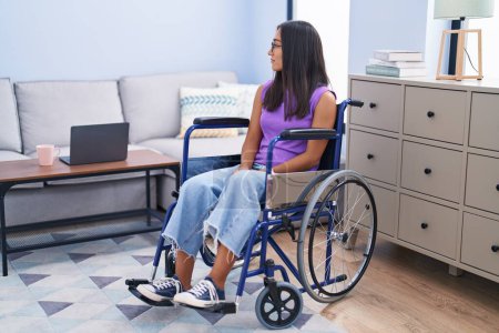 Foto de Young hispanic woman sitting on wheelchair at home looking to side, relax profile pose with natural face with confident smile. - Imagen libre de derechos