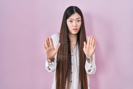 Foto de Mujer joven china de pie sobre fondo rosa alejando las palmas de las manos mostrando rechazo y negación con miedo y expresión repugnante. parada y prohibido. - Imagen libre de derechos