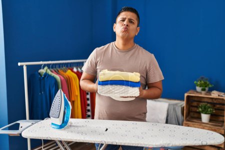 Photo for Hispanic young man holding folded laundry after ironing looking at the camera blowing a kiss being lovely and sexy. love expression. - Royalty Free Image