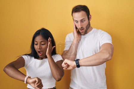 Foto de Interracial pareja de pie sobre el fondo amarillo mirando el reloj tiempo preocupado, miedo de llegar tarde - Imagen libre de derechos