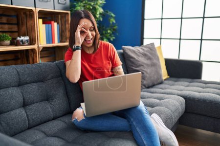 Foto de Young hispanic woman using laptop at home smiling happy doing ok sign with hand on eye looking through fingers - Imagen libre de derechos