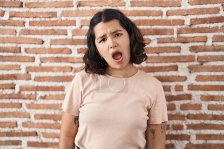 Téléchargez les photos : Jeune femme hispanique debout au-dessus du mur de briques dans le visage de choc, l'air sceptique et sarcastique, surpris avec la bouche ouverte - en image libre de droit