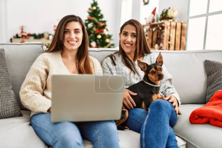 Photo for Two women using laptop sitting with dog by christmas tree at home - Royalty Free Image