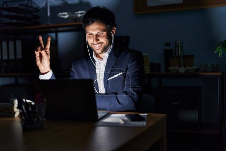 Photo for Handsome latin man working at the office at night smiling with happy face winking at the camera doing victory sign. number two. - Royalty Free Image
