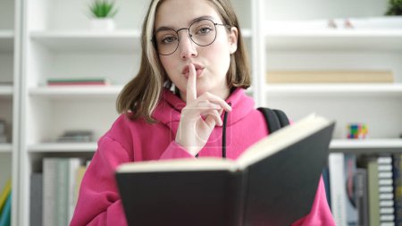 Téléchargez les photos : Young blonde woman student reading book doing silence gesture at library university - en image libre de droit
