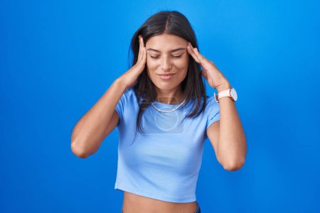 Photo for Brunette young woman standing over blue background with hand on head, headache because stress. suffering migraine. - Royalty Free Image