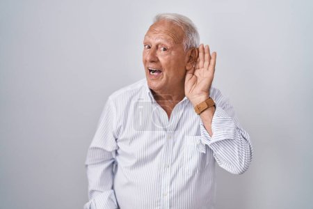 Téléchargez les photos : Senior man with grey hair standing over isolated background smiling with hand over ear listening an hearing to rumor or gossip. deafness concept. - en image libre de droit