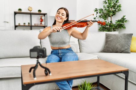 Photo for Young beautiful hispanic woman recording violin class sitting on sofa at home - Royalty Free Image