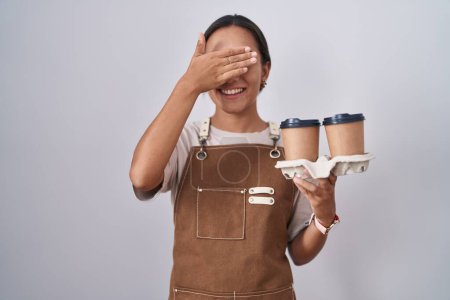 Foto de Mujer hispana joven vistiendo delantal de camarera profesional sosteniendo café sonriendo y riendo con la mano en la cara cubriendo los ojos para sorpresa. concepto ciego. - Imagen libre de derechos