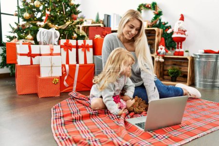 Photo for Mother and daughter using laptop sitting by christmas tree at home - Royalty Free Image