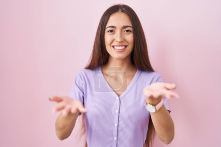 Foto de Joven mujer hispana con el pelo largo de pie sobre fondo rosa sonriente alegre ofreciendo las manos dando asistencia y aceptación. - Imagen libre de derechos