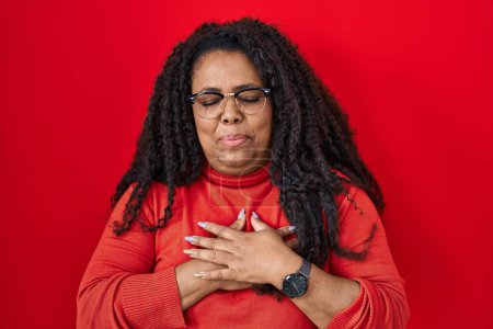 Photo for Plus size hispanic woman standing over red background smiling with hands on chest with closed eyes and grateful gesture on face. health concept. - Royalty Free Image