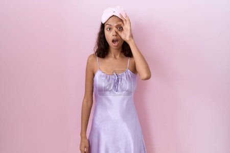 Téléchargez les photos : Young hispanic woman wearing sleep mask and nightgown doing ok gesture shocked with surprised face, eye looking through fingers. unbelieving expression. - en image libre de droit