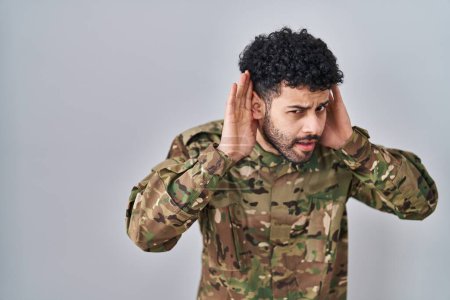 Foto de Hombre árabe vistiendo uniforme camuflaje del ejército tratando de escuchar ambas manos en el gesto de la oreja, curioso por chismes. problema auditivo, sordo - Imagen libre de derechos