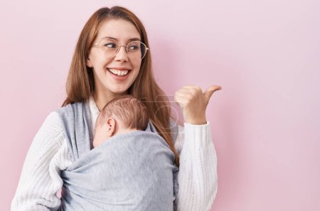 Foto de Joven mujer caucásica sosteniendo y llevando al bebé en un cabestrillo apuntando el pulgar hacia el lado sonriendo feliz con la boca abierta - Imagen libre de derechos