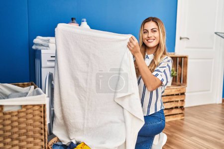 Foto de Joven mujer rubia sonriendo confiada sosteniendo la toalla en la lavandería - Imagen libre de derechos