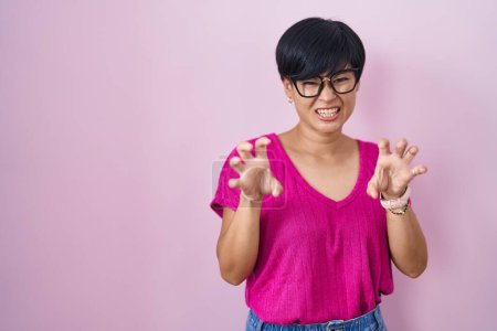 Téléchargez les photos : Jeune femme asiatique aux cheveux courts debout sur fond rose souriant drôle faisant griffe geste comme chat, expression agressive et sexy - en image libre de droit
