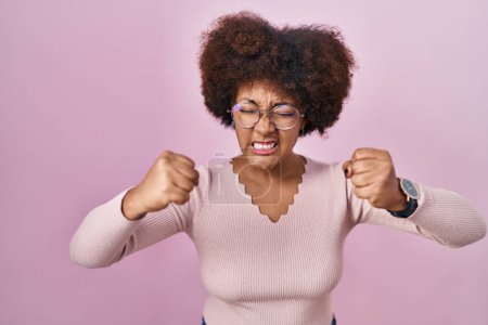 Photo for Young african american woman standing over pink background angry and mad raising fists frustrated and furious while shouting with anger. rage and aggressive concept. - Royalty Free Image