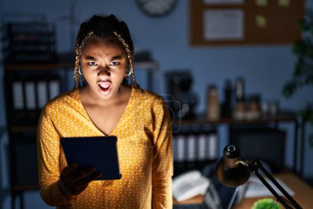 Téléchargez les photos : African american woman with braids working at the office at night with tablet angry and mad screaming frustrated and furious, shouting with anger. rage and aggressive concept. - en image libre de droit