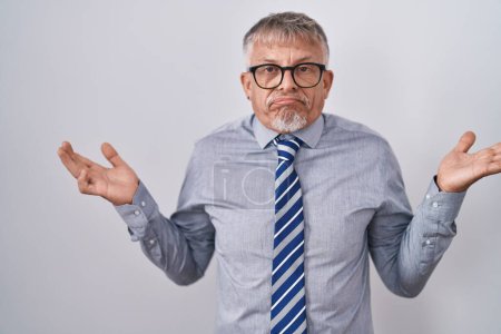 Téléchargez les photos : Hispanic business man with grey hair wearing glasses clueless and confused expression with arms and hands raised. doubt concept. - en image libre de droit