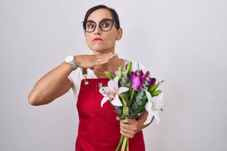 Foto de Mujer morena de mediana edad vistiendo delantal trabajando en floristería sosteniendo ramo degollando con la mano como cuchillo, amenazan la agresión con violencia furiosa - Imagen libre de derechos